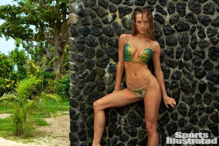 Hannah Ferguson poses in front of a stone wall wearing a rainbow-colored metallic swimsuit.